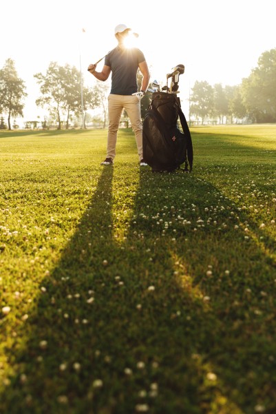 Young man in cap holding golf club while standing on a green course with black golf bag